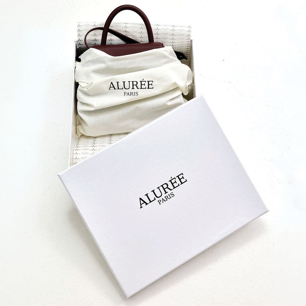 An ALURÉE PARIS handbag shown inside its white presentation box, with a white dust bag and a dark brown leather handle visible. The white branded box is placed in the foreground on a clean white surface.